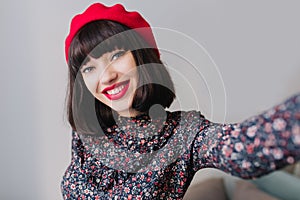 Gorgeous stylish brunette girl wearing retro dress with floral print smiling widely while making selfie. Close-up