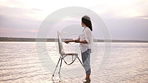 Gorgeous view of a young curly brunette standing in the water with easel and draing her the surrounding landscape