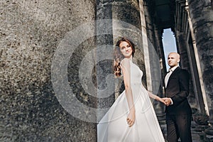 Gorgeous wedding couple embracing in sunlight near old castle in beautiful park. Stylish beautiful bride and groom gently hugging