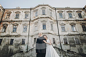 Gorgeous wedding couple embracing in sunlight near old castle in beautiful park. Stylish beautiful bride and groom gently hugging