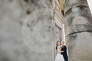 Gorgeous wedding couple embracing in sunlight near old castle in beautiful park. Stylish beautiful bride and groom gently kissing