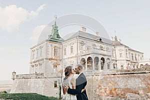 Gorgeous wedding couple embracing in sunlight near old castle in beautiful park. Stylish beautiful bride and groom gently kissing