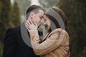 Gorgeous wedding couple kissing in winter snowy park. stylish bride in coat and  groom embracing under green trees in winter