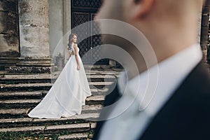 Gorgeous wedding couple posing on stone stairs near old castle in park. Stylish beautiful bride in amazing gown looking at groom