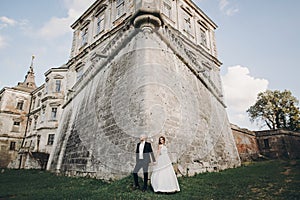 Gorgeous wedding couple posing in sunlight near old castle in beautiful park. Stylish beautiful bride and groom holding hands on