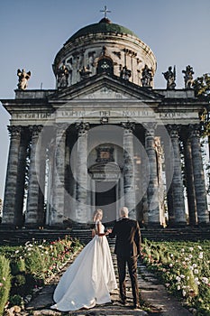 Gorgeous wedding couple walking near old castle in sunny beautiful park. Stylish beautiful bride and groom posing on background of