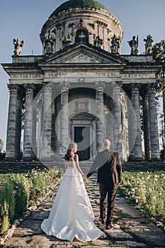 Gorgeous wedding couple walking near old castle in sunny beautiful park. Stylish beautiful bride and groom posing on background of