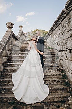 Gorgeous wedding couple walking on stone stairs near old castle in park. Stylish beautiful bride in amazing gown and  groom posing