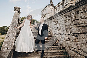 Gorgeous wedding couple walking on stone stairs near old castle in park. Stylish beautiful bride in amazing gown and  groom posing