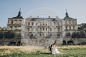 Gorgeous wedding couple walking in sunlight near old castle in beautiful park. Stylish beautiful bride and groom holding hands on