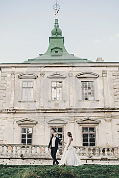 Gorgeous wedding couple walking in sunlight near old castle in beautiful park. Stylish beautiful bride and groom posing on