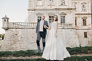 Gorgeous wedding couple walking in sunlight near old castle in beautiful park. Stylish beautiful bride and groom posing on