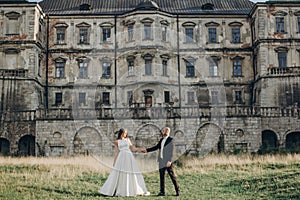 Gorgeous wedding couple walking in sunlight near old castle in beautiful park. Stylish beautiful bride and groom holding hands on