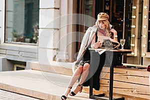 Gorgeous woman in black dress resting in outdoor cafe and reading newspaper. Elegant girl in brown coat and hat sitting