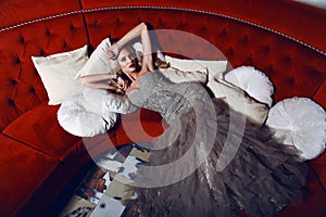 Gorgeous woman with blond hair in elegant dress lying on red divan photo