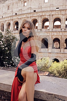 gorgeous woman with blond hair in luxurious red evening dress, gloves and jewelry posing near the Colosseum in Rome