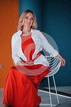 Gorgeous woman. Blonde in festive red dress is in the room with orange and blue colored walls and white chair