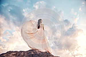 Gorgeous woman brunette in the mountains at sunset and blue sky with clouds. The woman looks into the distance in a long white