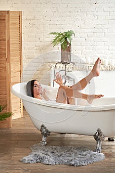 A gorgeous woman is enjoying herself in a white bathtub, in a bright room with a large window