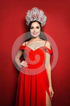 Gorgeous woman fashion model wearing red dress and diamond crown on red background