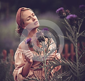 Gorgeous woman in off-shoulder summer clothes stands in the middle of field, touches thistle thorns, feeling aroma