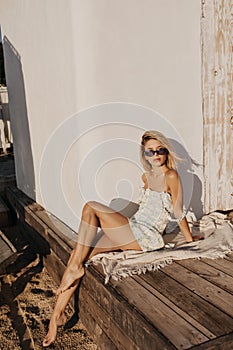 Gorgeous young blonde sits on wooden floor sunbathing on beach.