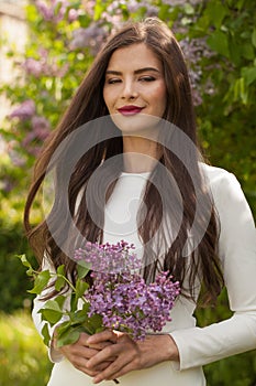 Gorgeous young brunette woman with long wavy hair wearing white dress holding bouquet of spring flowers