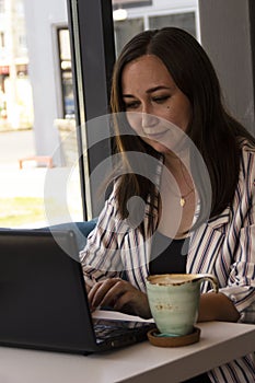 Gorgeous young business woman working remotely while sitting in front of an open laptop computer in the interior of a modern cafe