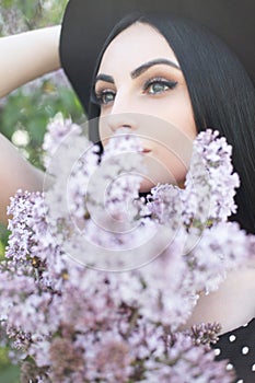 Gorgeous young woman with amazing eyes with clean skin and lilac bouquet flowers on nature. Attractive girl in elegant hat covers