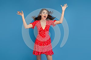 Gorgeous young woman with bright smile in red casual dress looking at camera with excited face with hands up over blue