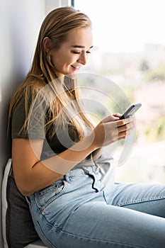 Gorgeous young woman checking email and sending sms message on mobile phone sitting on windowsill