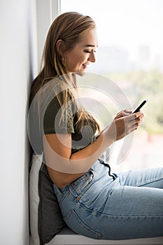 Gorgeous young woman checking email and sending sms message on mobile phone sitting on windowsill