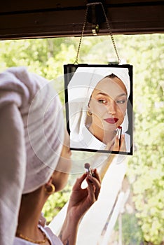 Gorgeous young woman doing her makeup while sitting in front of the mirror by the window