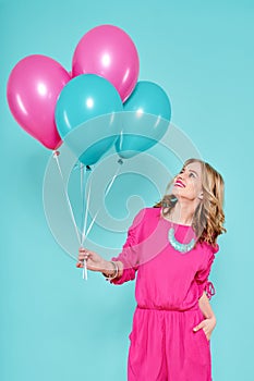 Gorgeous young woman in party summer dress holding bunch of colourful balloons, isolated over pastel blue colored background.