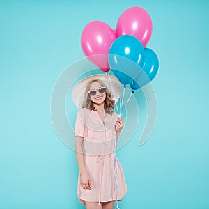 Gorgeous young woman in party summer dress and straw hat holding bunch of colourful balloons, isolated over pastel blue.