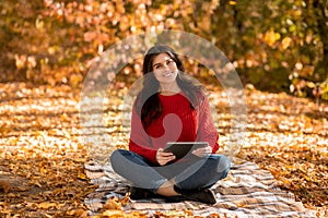 Gorgeous young woman in red knitted sweater using tablet computer at autumn park