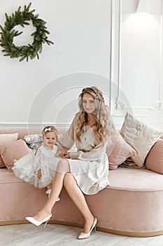 Gorgeous young woman in stylish dress posing with her little daughter in fluffy dress in Christmas decorated interior