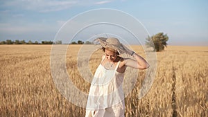 Gorgeous young woman in white dress runnung in the wheat field. Smiling, happy female running and holds her straw hat on