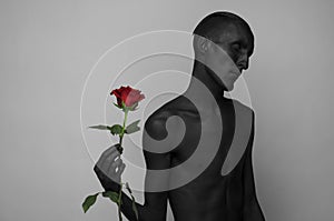 Gothic and Halloween theme: a man with black skin holding a red rose, black death on a gray background in studio