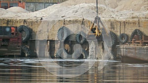 The grab bucket of the excavator rises from the river with splashes of water in the port against the background of an