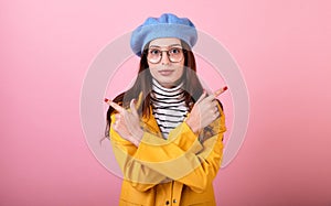 Graceful girl in a stylish beret hat and a yellow raincoat in glasses points her fingers in different directions on a pink