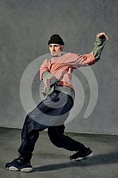 Graceful male with tattooed face, beard. Dressed in colorful jumper, black pants and sneakers. Dancing on gray