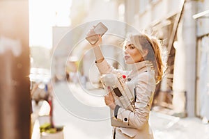 Graceful slim girl with blonde ponytail waving hand with smartphone and looking at the road. Joyful lady in light-brown
