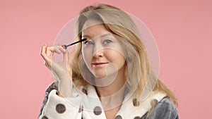 Graceful Woman Applying Mascara to Eyelashes, Focused and Elegant Against a Simple Pink Background