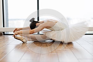 Graceful woman ballet dancer stretching while sitting on wooden floor.