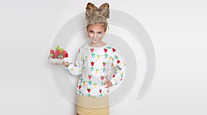 Gracious little girl with blond hair and blue eyes standing on a white background wearing a sweatshirt and a strawberry