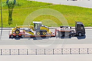Grader tractor on transportation trailer truck with long trailer platform on the highway in the city