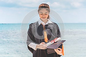 Graduated woman in cap and gown holding certificated celebrating in Commencement day with blue sky on the beach