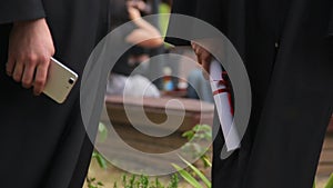 Graduates holding smartphones and diplomas, waiting for graduation ceremony