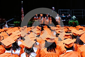Graduates sitting in orange caps and gowns with stage and speakers in background II